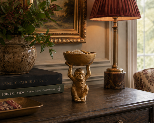 A decorative gold-toned monkey figurine holding a bowl of popcorn sits on a dark wooden sideboard in a styled room setting with a lamp, books, and a plant in the background.