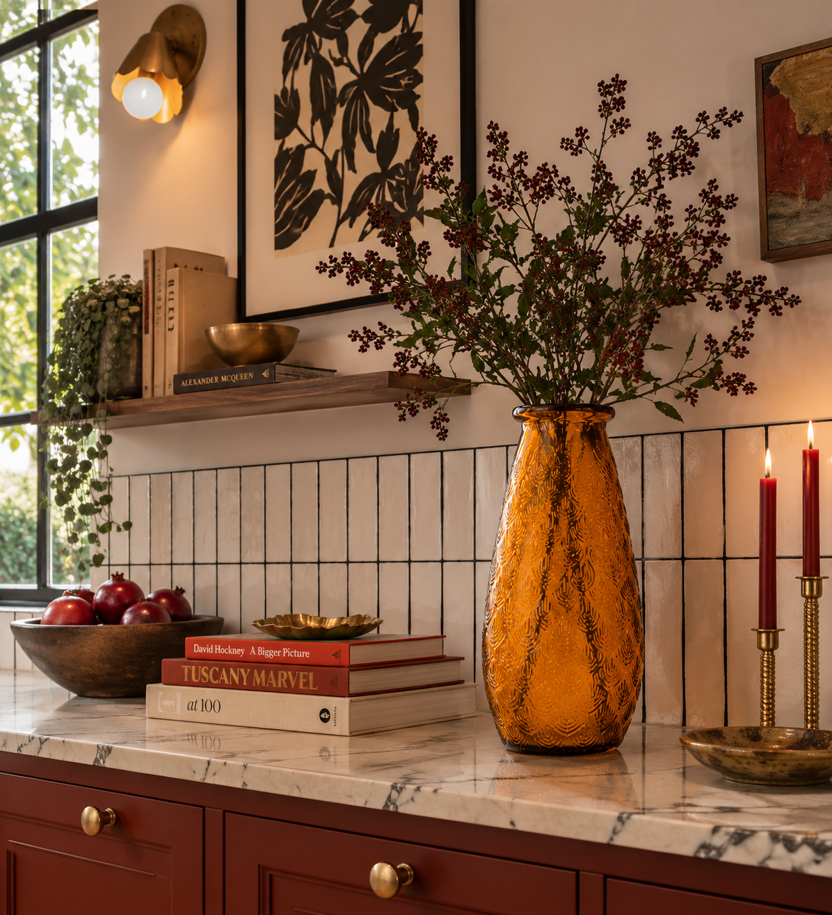 A warm, styled interior scene featuring a textured amber-orange glass vase filled with dried purple-brown flowers on a marble countertop. The setting includes a white subway tile backsplash, red wooden cabinets, a bowl of red apples, books, and lit red taper candles.