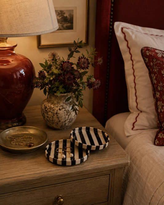 A styled bedroom nightstand scene with a red ceramic table lamp, a vase of dark flowers, a small tray with jewelry, and two round black-and-white striped jewelry rings trays in the foreground beside a bed with cream bedding and red pillows.