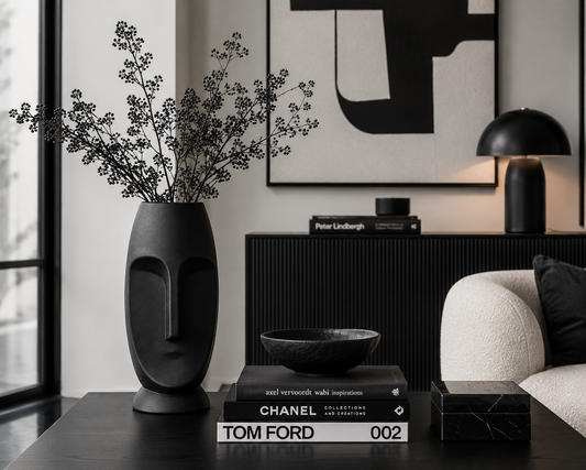 A black ceramic face-shaped vase with dried botanical stems, styled on a dark table in a modern living room setting next to a white armchair and a black sideboard.